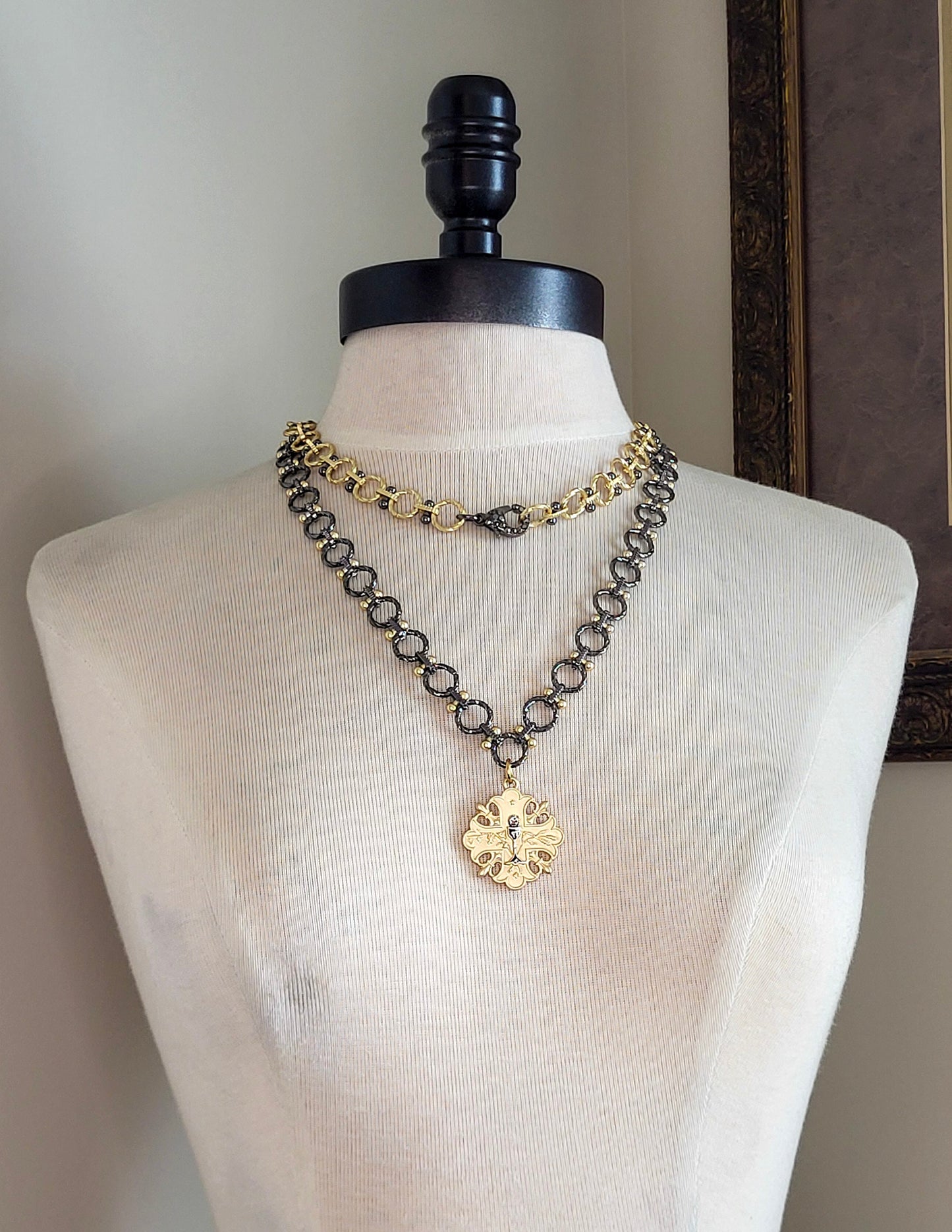 A 20 inch gun metal and gold chain necklace with a gold pendant. The pendant is a cross design with a floral pattern and a small gun metal chalice in the center.