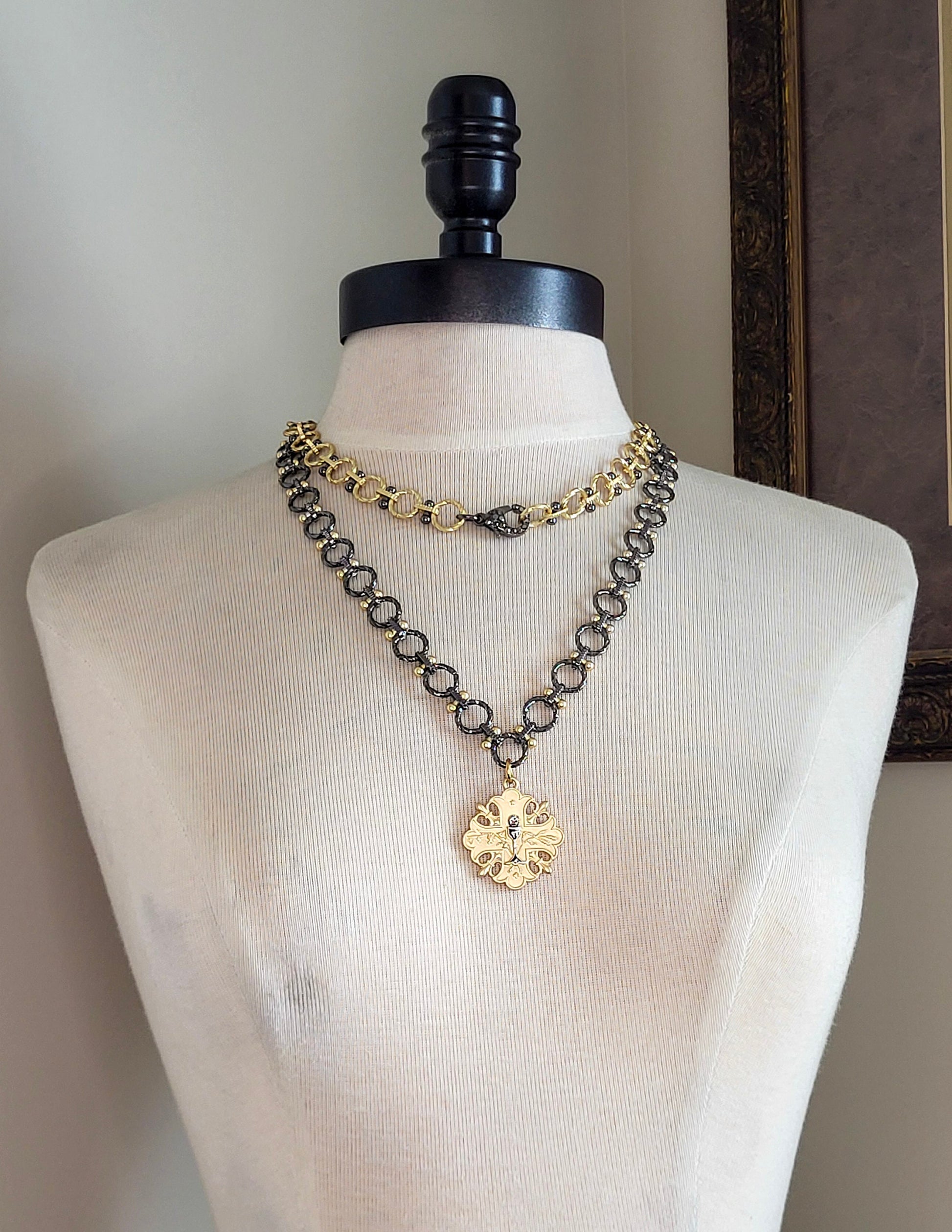 A 20 inch gun metal and gold chain necklace with a gold pendant. The pendant is a cross design with a floral pattern and a small gun metal chalice in the center.
