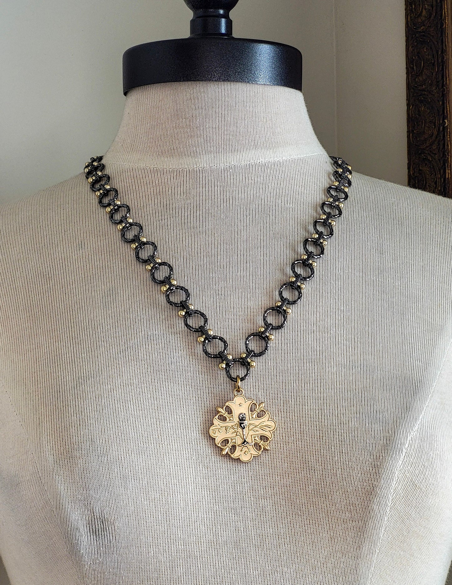 A gun metal and gold chain necklace with a gold pendant. The pendant is a cross design with a floral pattern and a small gun metal chalice in the center.