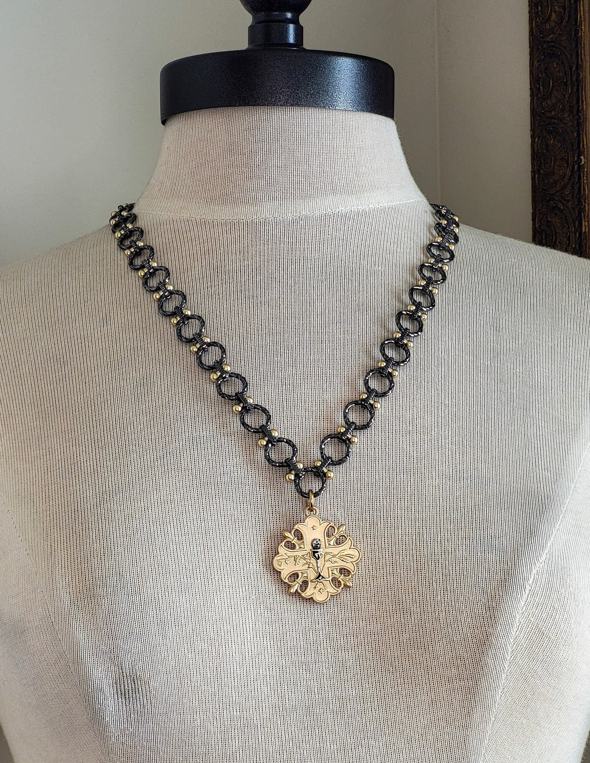 A gun metal and gold chain necklace with a gold pendant. The pendant is a cross design with a floral pattern and a small gun metal chalice in the center.