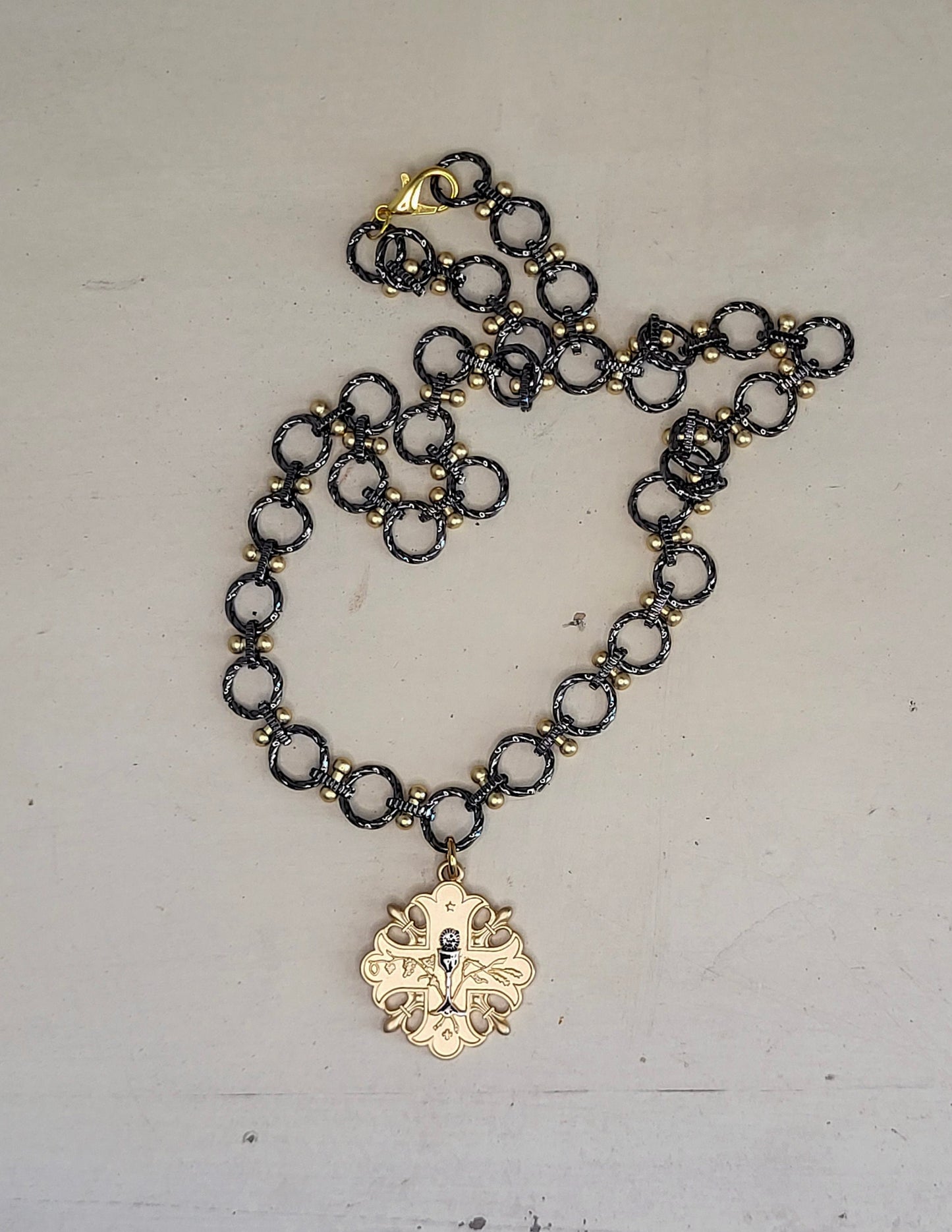 A 20 inch gun metal and gold chain necklace with a gold pendant. The pendant is a cross design with a floral pattern and a small gun metal chalice in the center.