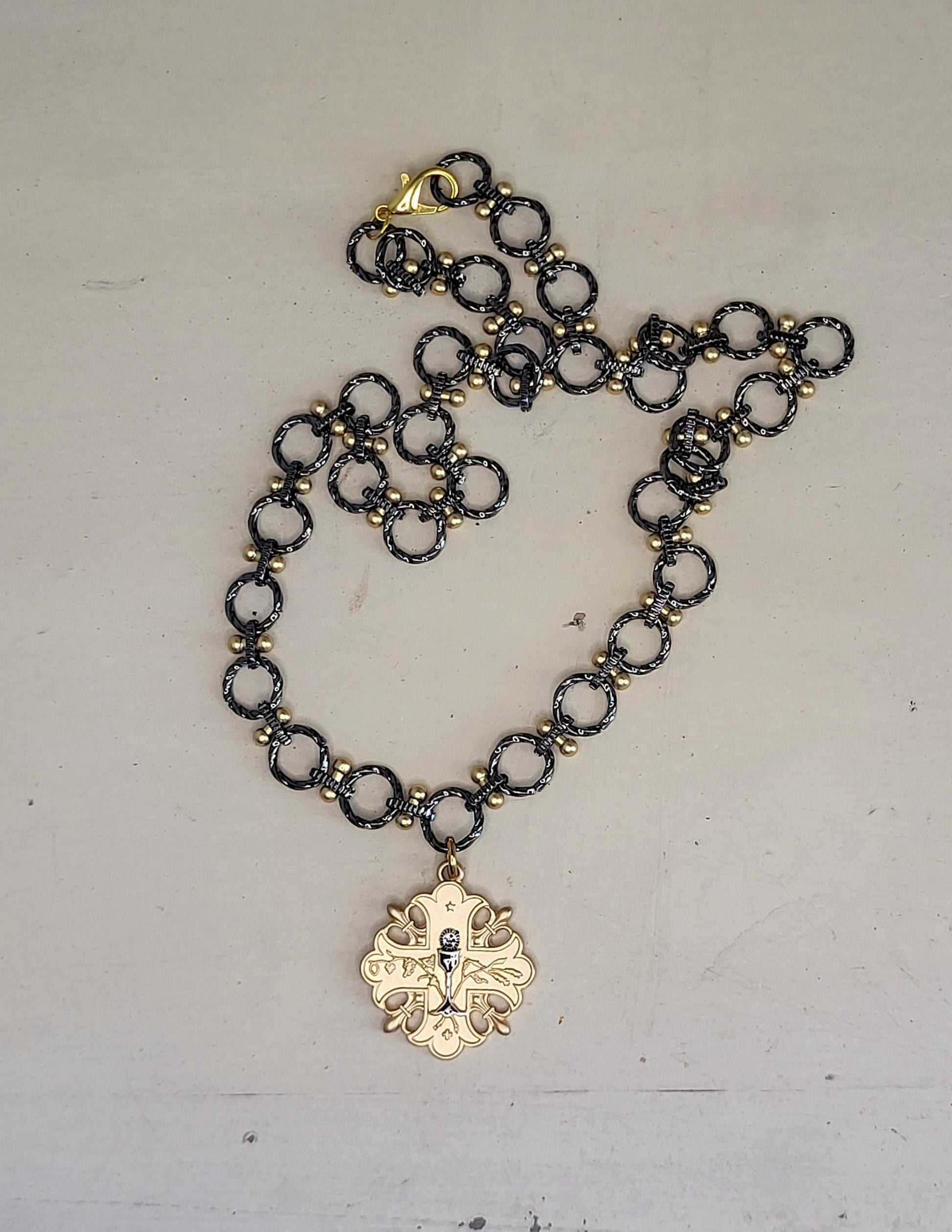 A 20 inch gun metal and gold chain necklace with a gold pendant. The pendant is a cross design with a floral pattern and a small gun metal chalice in the center.