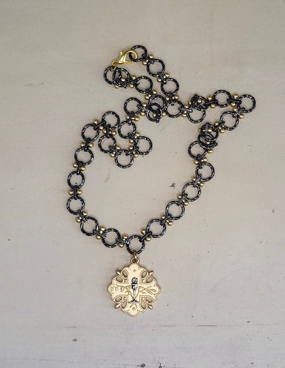 A 20 inch gun metal and gold chain necklace with a gold pendant. The pendant is a cross design with a floral pattern and a small gun metal chalice in the center.
