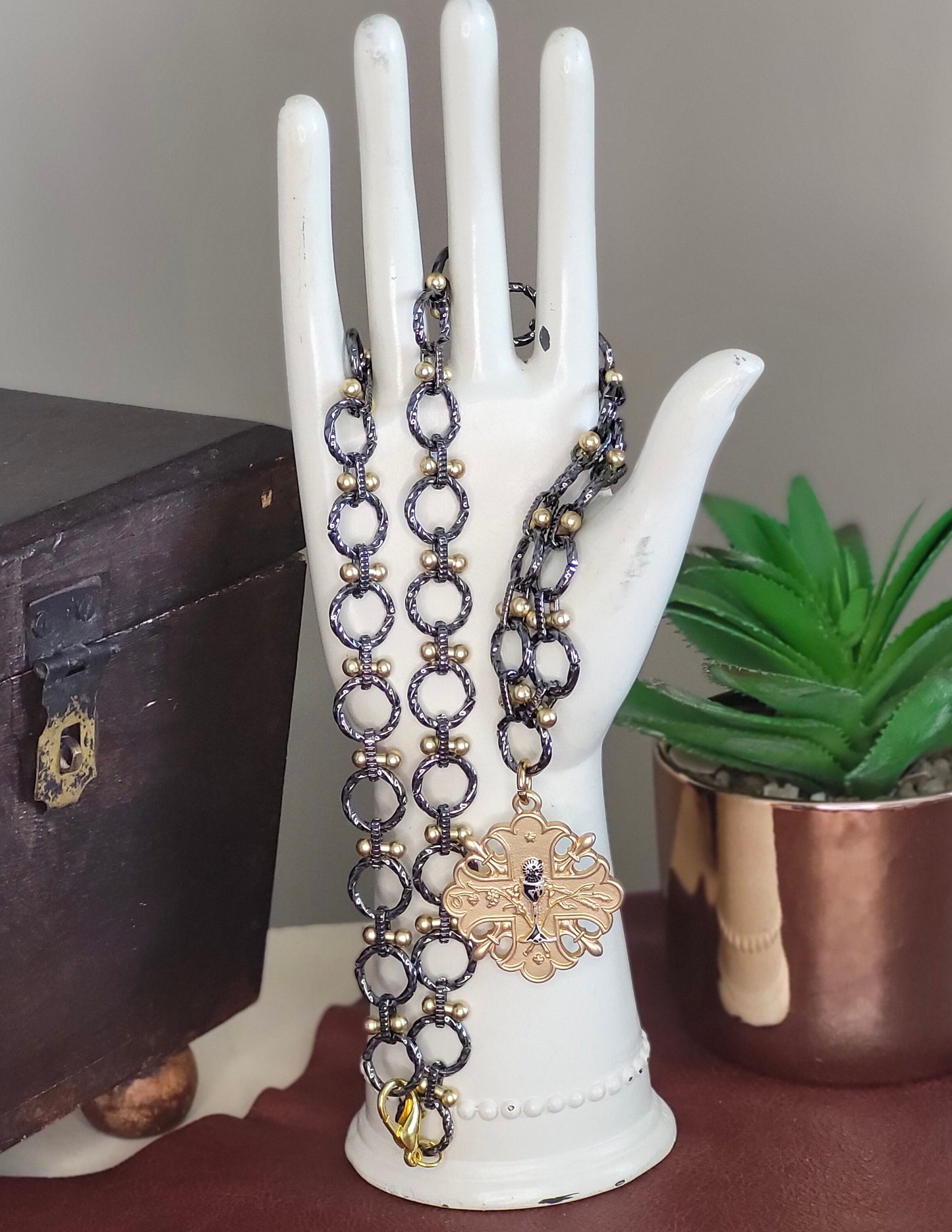 May include: A gun metal and gold chain necklace with a gold pendant featuring a cross design. The pendant has a detailed design with a small chalice in the center.
