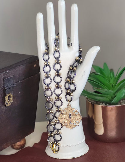 May include: A gun metal and gold chain necklace with a gold pendant featuring a cross design. The pendant has a detailed design with a small chalice in the center.
